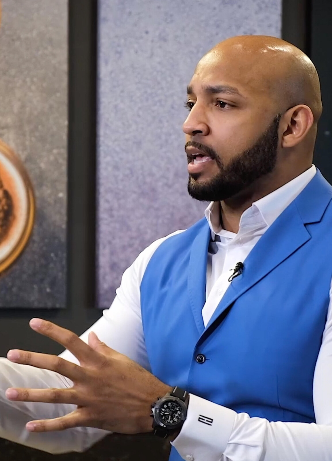 Roy Hall wearing a blue vest and speaking during an interview